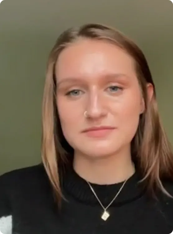 Portrait of a young woman with light brown hair wearing a black sweater and a necklace with a square pendant.