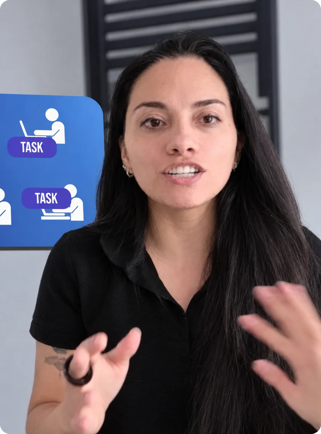 Woman with long dark hair speaking and gesturing with her hands in front of a screen displaying icons of people working on laptops with the word 'TASK'.