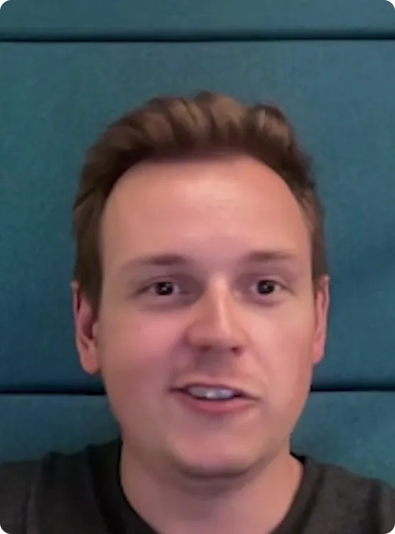 Close-up of a young man with short light brown hair speaking in front of a teal padded wall.