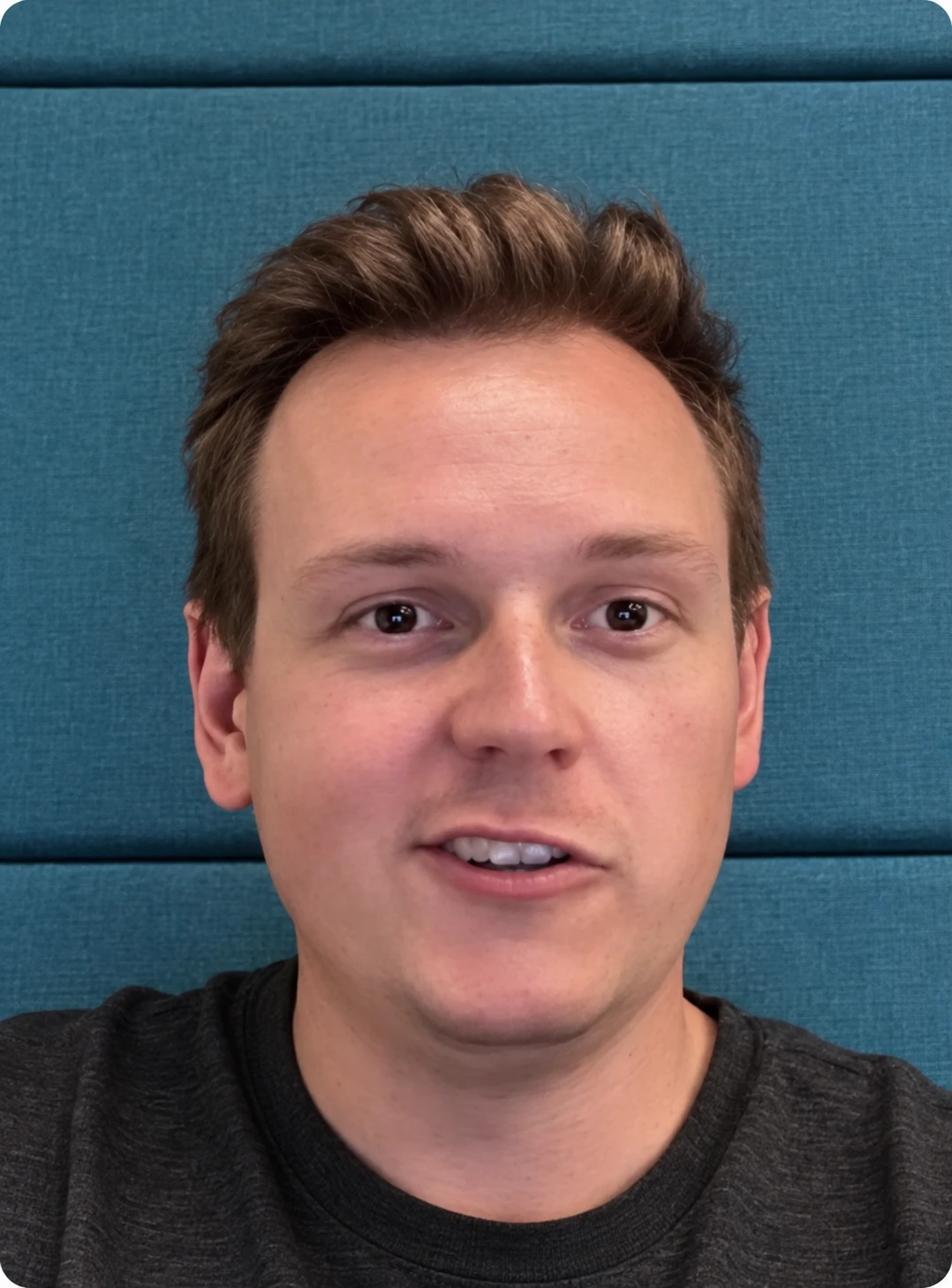 Close-up of a young man with short light brown hair speaking in front of a teal padded wall.
