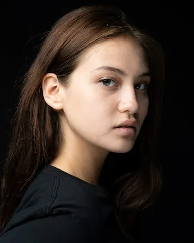 Portrait of a young woman with light skin and brown hair against a black background, looking slightly to the side.