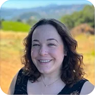 Smiling woman with curly brown hair standing outdoors with blurred fields and hills in the background.