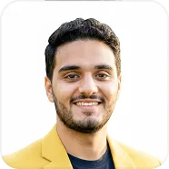 Smiling man with short curly black hair and beard wearing a yellow blazer and black shirt, against a white background.